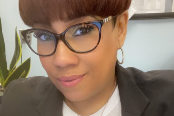 A woman with short hair and glasses poses for a photo wearing a blazer and white shirt.