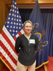 A woman stands in front of the United States and Pennsylvania state flags.