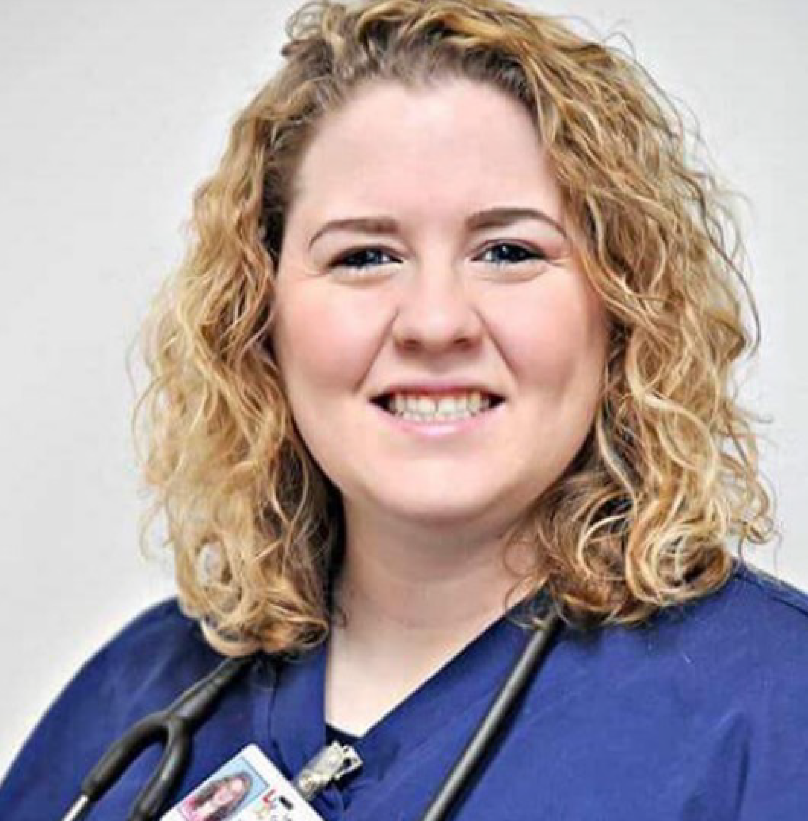 Headshot of a woman with blonde curly hair wearing blue scrubs.