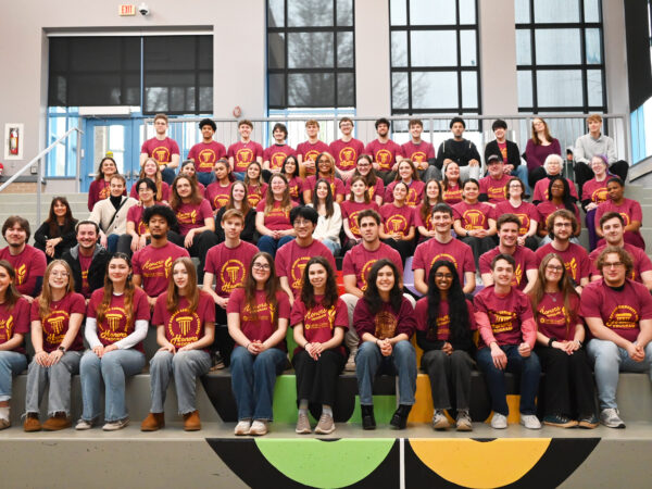 A large group of approximately 50 college students and faculty members posing for a formal group portrait on a wide set of indoor stairs. Most individuals are wearing matching maroon t-shirts that feature a gold torch logo and the words "Honors Program" and "Lehigh Carbon Community College."