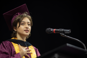 A woman in commencement regalia holds her right hand over her heart as she speaks at a podium microphone.