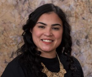 Professional portrait of a woman with long black hair wearing a black shirt and gold necklace.