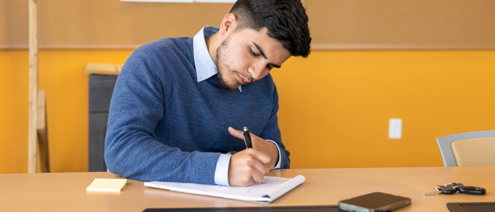 A young man in a dress shirt and pull over sweater writes at a table.