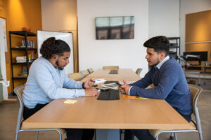 Two men sit across from each at a long work table.