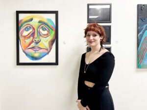 A student with red hair and wearing all black stands in front of a multicolored piece of artwork that is a the face of a person, eyes upturned. 