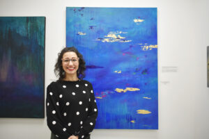 A woman in a black and white polka dot sweater stands next to a large painting featuring the color blue and some white that appear to be clouds.
