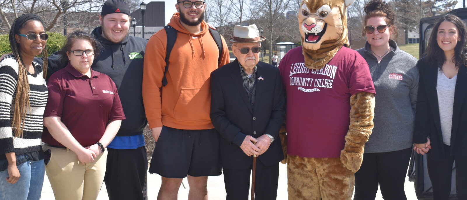 A man wearing a hat, sunglasses and carrying a cane poses in the middle of a group of students and Clawrence the Cougar mascot.