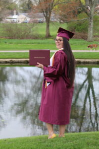 A graduate wearing commencement regalia holds their LCCC diploma cover in front of a pond.