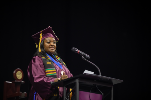 A woman in commencement regalia stands at a podium microphone.