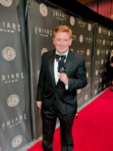 A man in a tuxedo holds a microphone while standing on a red carpet in front of a wall that says "Friars Club"