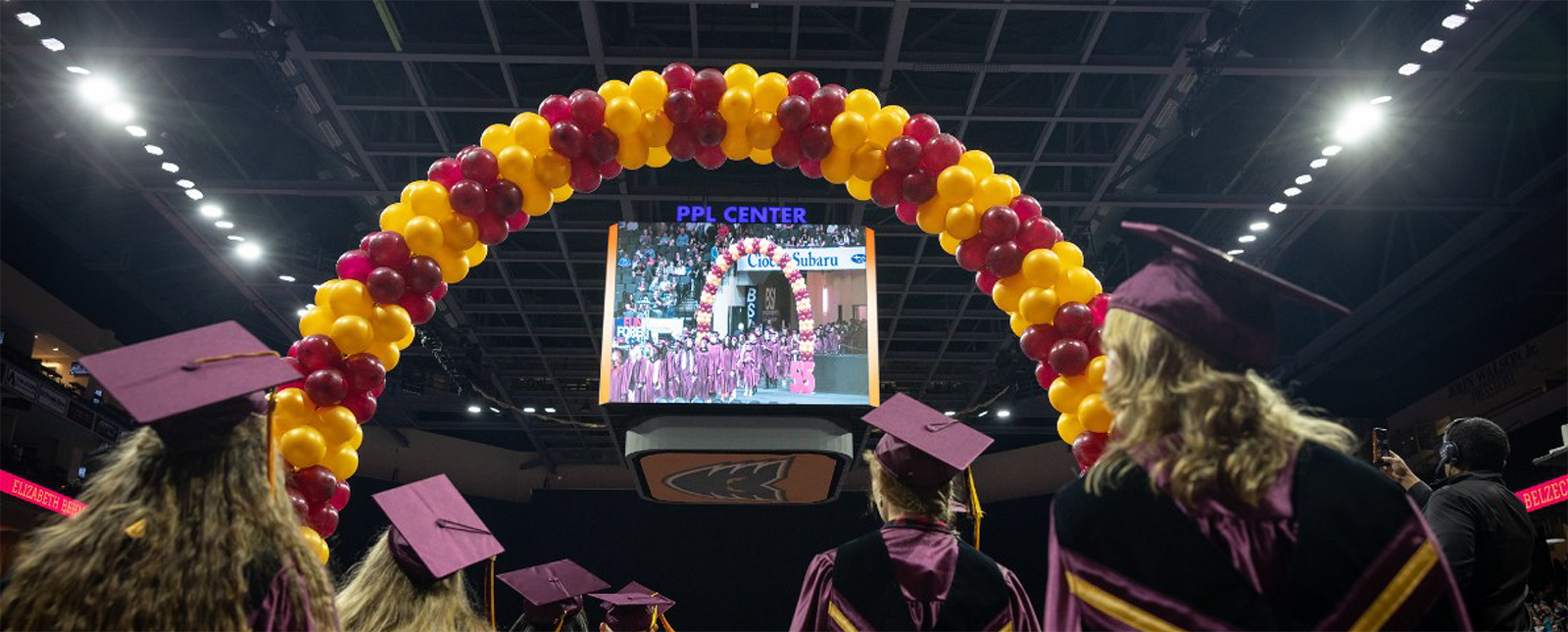 The backs of graduates can be seen as they look towards a large screen in an arena through a maroon and gold balloon arch.