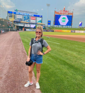 A woman wearing a gray t-shirt and jean shorts stands on the field at a baseball stadium while holding a camera.