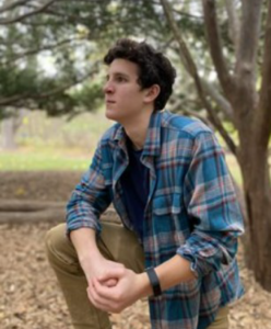 A young man in a blue plaid shirt and khakis kneels on one knee in a park and looks off camera to the left.