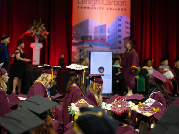 An image showing a commencement ceremony at Lehigh Carbon Community College (LCCC). Graduates wearing maroon academic gowns and caps are seated facing a stage with red drapes and a backdrop bearing the college's name.