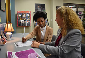 Image shows a student sitting at a table while writing and speaking with a staff member.