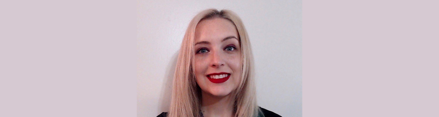 A blonde woman wearing red lipstick poses for a photo against a white wall.