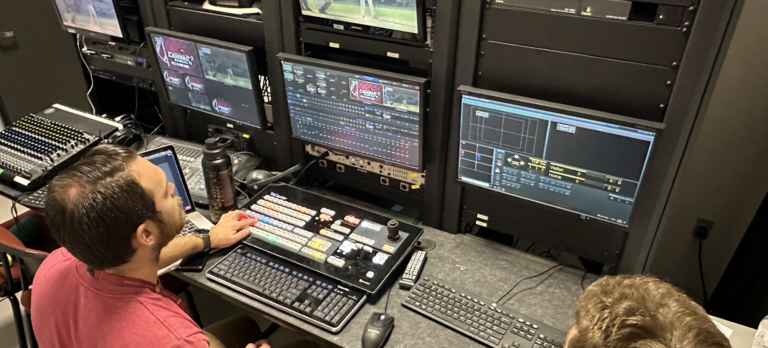 An instructor sits at a bank of TV monitors next to a student.